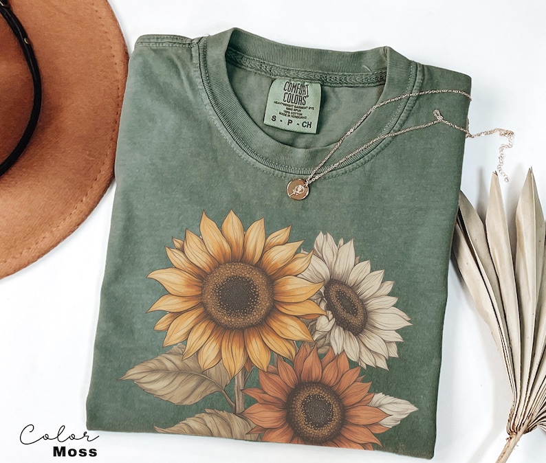 May include: A green t-shirt featuring a sunflower design. The shirt has a distressed look and is made from a soft, comfortable fabric. The text "COMFORT COLORS" is visible on the tag.