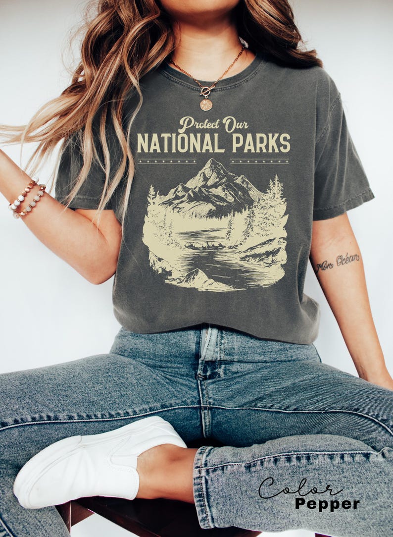 May include: A charcoal grey t-shirt with a vintage-style graphic of a mountain range, a lake, and trees. The text "Protect Our National Parks" is printed in a cream color on the shirt.