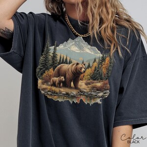May include: Black t-shirt featuring a detailed illustration of a bear and cub in a mountain landscape. The design includes a lake, trees, and a mountain range in the background. The word "Color" and "BLACK" are printed on the bottom right.