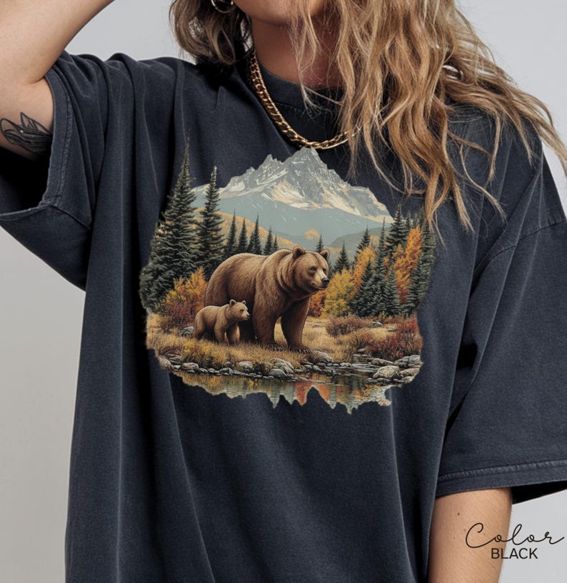 May include: Black t-shirt featuring a detailed illustration of a bear and cub in a mountain landscape. The design includes a lake, trees, and a mountain range in the background. The word "Color" and "BLACK" are printed on the bottom right.
