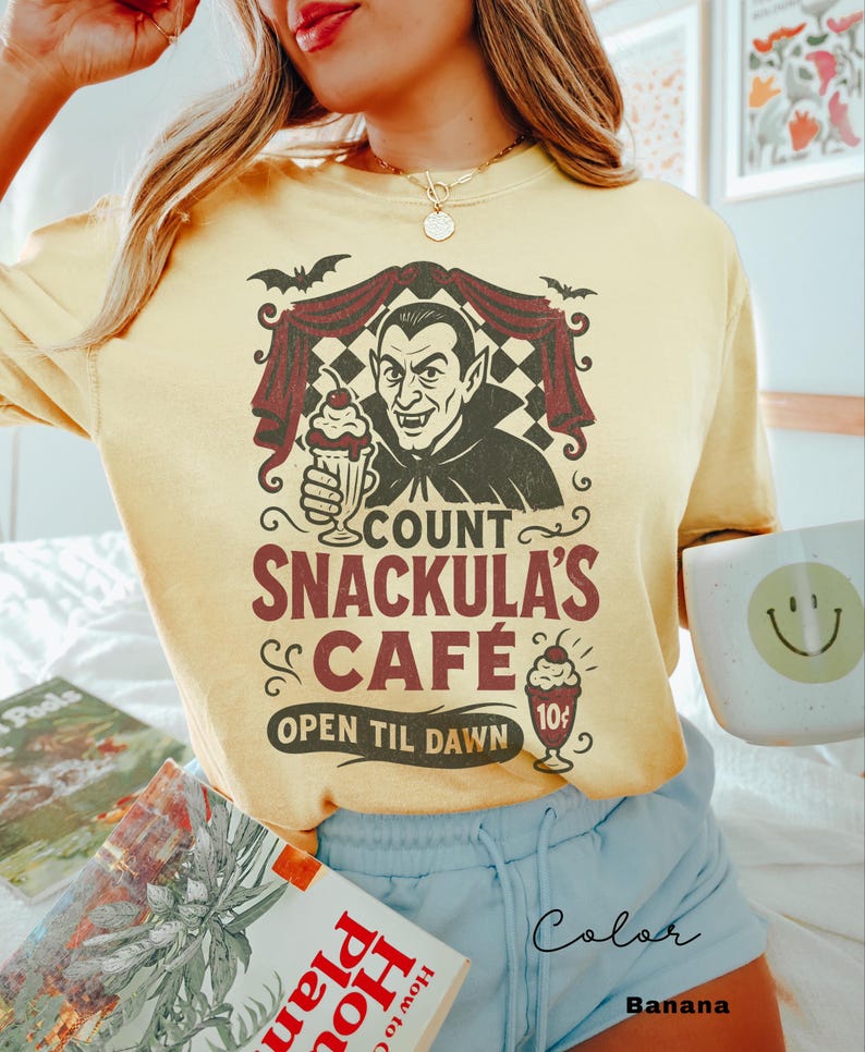 May include: A light yellow t-shirt with a graphic of Count Snackula holding a milkshake. The design includes bats, a checkered background, and the text "Count Snackula's Cafe, Open Til Dawn". The shirt is paired with light blue shorts.