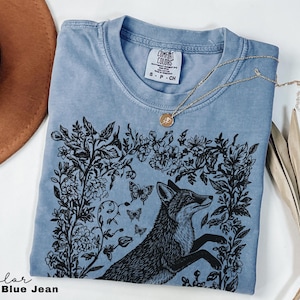 Pu&ograve; includere: T-shirt blu jeans con grafica di volpe nera circondata da disegni floreali e farfalle. La maglietta ha girocollo e maniche corte. Una collana d'oro con un ciondolo &egrave; drappeggiata sulla maglietta. Il testo "Color Blue Jean" &egrave; in basso a sinistra.