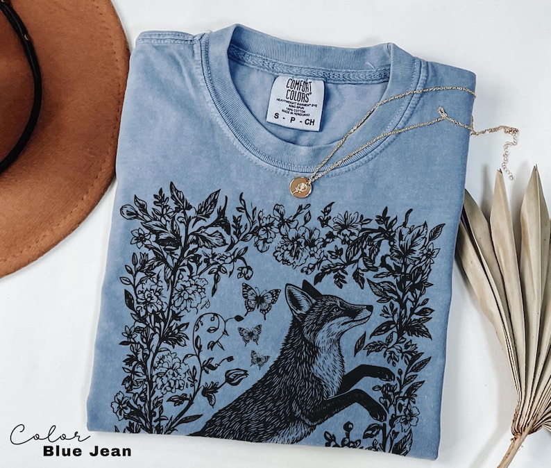 Pu&ograve; includere: T-shirt blu jeans con grafica di volpe nera circondata da disegni floreali e farfalle. La maglietta ha girocollo e maniche corte. Una collana d'oro con un ciondolo &egrave; drappeggiata sulla maglietta. Il testo "Color Blue Jean" &egrave; in basso a sinistra.