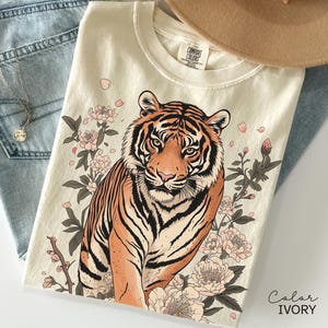 May include: Ivory-colored t-shirt featuring a detailed illustration of a tiger surrounded by pink flowers and foliage. The tiger has orange and black stripes. The shirt is folded on top of blue jeans. The text "Color IVORY" is visible.