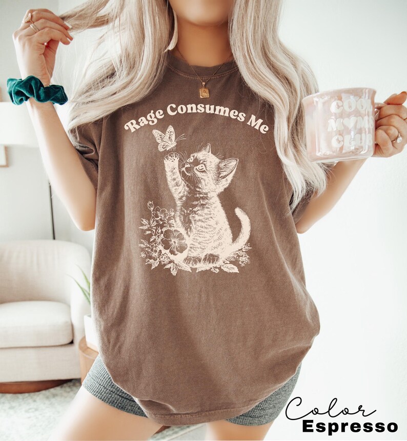 May include: A brown t-shirt with a white graphic of a kitten reaching for a butterfly. The text "Rage Consumes Me" is printed on the shirt. The shirt is shown on a model wearing it.