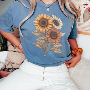 May include: A woman wearing a blue jean t-shirt with a sunflower graphic. The t-shirt has a distressed look and the sunflowers are in shades of yellow, orange, and brown. The woman is wearing white jeans and has her hair down.