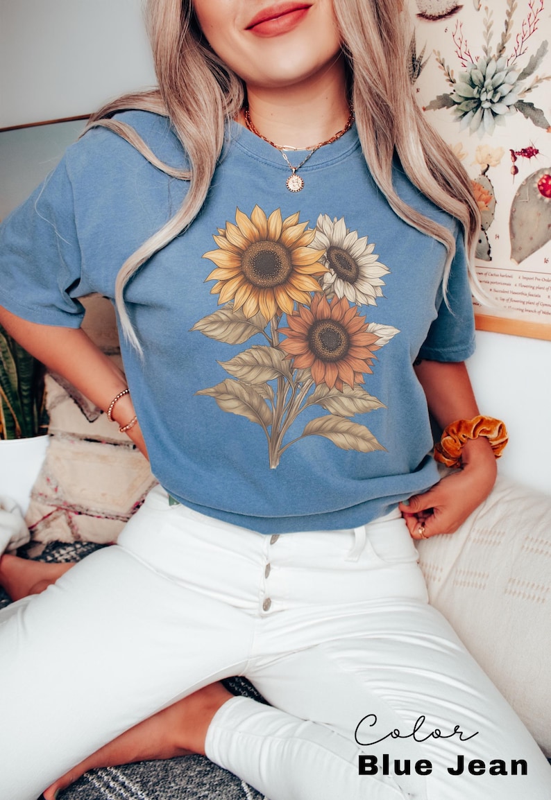 May include: A woman wearing a blue jean t-shirt with a sunflower graphic. The t-shirt has a distressed look and the sunflowers are in shades of yellow, orange, and brown. The woman is wearing white jeans and has her hair down.