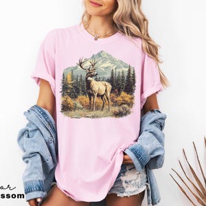 May include: A light pink t-shirt featuring a detailed illustration of a deer with large antlers standing in a forest scene with a mountain backdrop. The text "Color Blossom" is at the bottom.