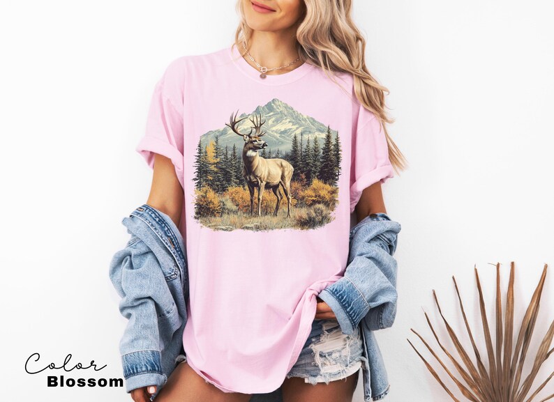May include: A light pink t-shirt featuring a detailed illustration of a deer with large antlers standing in a forest scene with a mountain backdrop. The text "Color Blossom" is at the bottom.