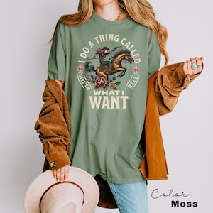 May include: A green t-shirt featuring a vintage-style illustration of a woman riding a horse. The text on the shirt reads "I do a thing called give 'em hell what I want".
