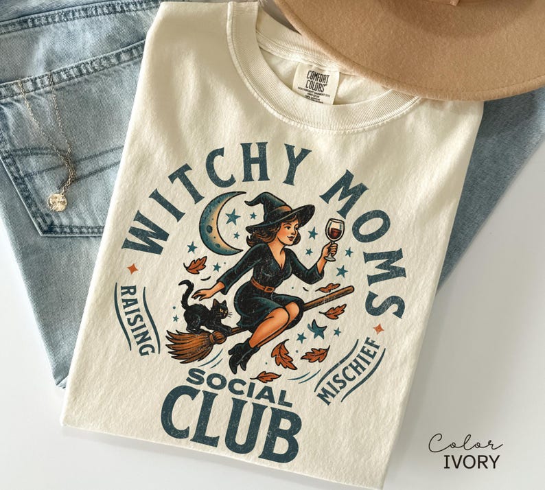 Puede incluir: Camiseta color marfil con un dise&ntilde;o de una bruja en una escoba, sosteniendo una copa de vino, con un gato negro. El texto dice "Witchy Moms Social Club, Raising Mischief". La camiseta est&aacute; doblada sobre unos vaqueros azules.