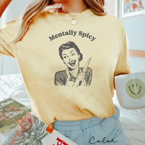 Puede incluir: Una camiseta amarilla con el texto "Mentally Spicy" y una ilustraci&oacute;n retro de una mujer sosteniendo un cuchillo. La mujer sonr&iacute;e y lleva un vestido y un delantal.