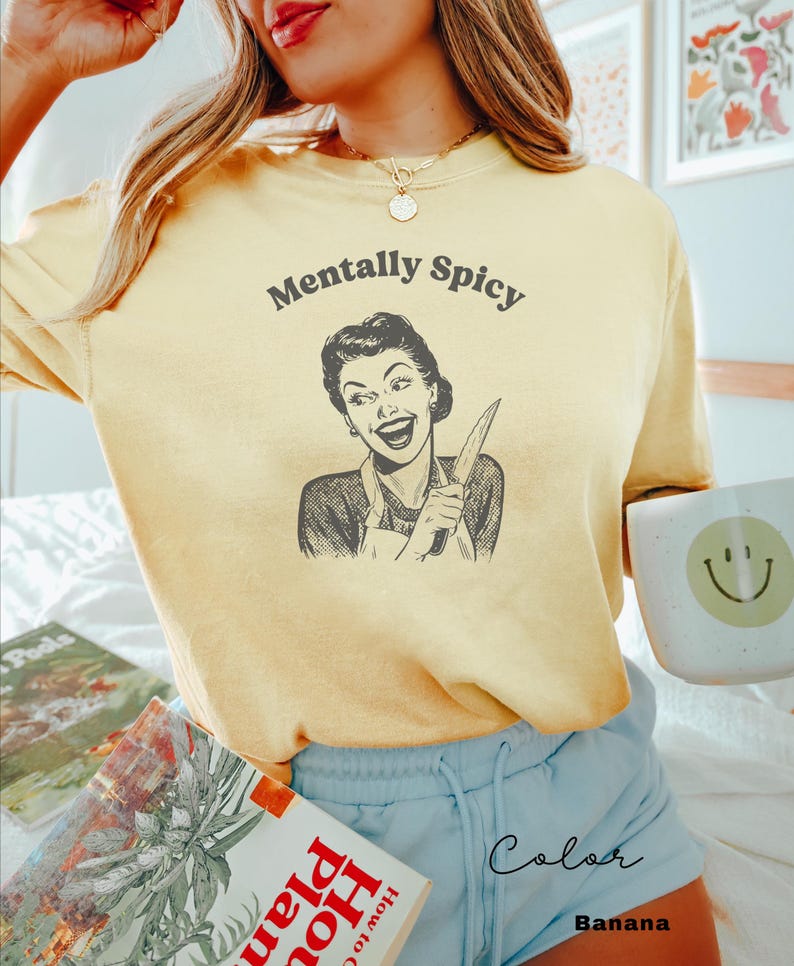 Puede incluir: Una camiseta amarilla con el texto "Mentally Spicy" y una ilustraci&oacute;n retro de una mujer sosteniendo un cuchillo. La mujer sonr&iacute;e y lleva un vestido y un delantal.