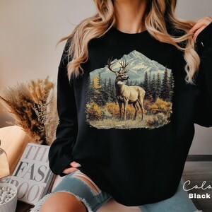 May include: Black sweatshirt featuring a detailed illustration of a deer in a mountain landscape. The graphic includes a deer with large antlers, trees, and a snow-capped mountain. The word "Color" and "Black" are written on the bottom right.