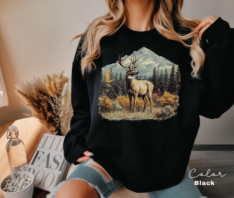 May include: Black sweatshirt featuring a detailed illustration of a deer in a mountain landscape. The graphic includes a deer with large antlers, trees, and a snow-capped mountain. The word "Color" and "Black" are written on the bottom right.