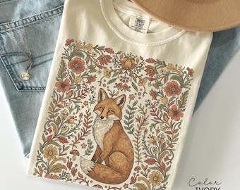 May include: Ivory-colored t-shirt featuring a detailed illustration of a fox surrounded by a floral design in shades of brown, orange, and green. The shirt is folded, with a hat and jeans visible in the background.