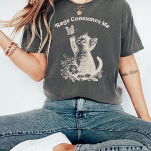 May include: A dark gray t-shirt with the text "Rage Consumes Me" printed on the front. A cartoon kitten is reaching up to catch a butterfly. The kitten is surrounded by flowers.