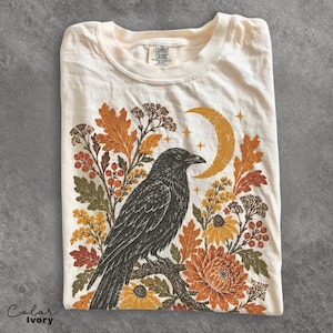 Puede incluir: Una camiseta color marfil con un estampado gráfico de un cuervo negro posado en una rama. El diseño incluye hojas de otoño naranjas y amarillas, bayas, flores y una luna creciente con estrellas pequeñas. La etiqueta del cuello dice Comfort Colors.
