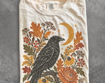May include: An ivory t-shirt featuring a graphic print of a black crow perched on a branch. The design includes orange and yellow autumn leaves, berries, flowers, and a crescent moon with small stars. The neck label reads Comfort Colors.
