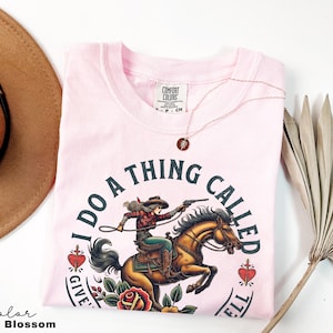 May include: Pink t-shirt featuring a graphic of a woman riding a horse with the text "I do a thing called give 'em hell".