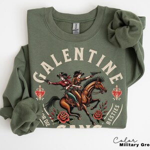 May include: Military green sweatshirt with a graphic featuring two cowgirls riding a horse, holding guns, and the text "GALENTINE BESTIES FOR DIE". The design includes roses and a heart.