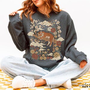May include: Dark heather gray sweatshirt featuring a tiger resting on a branch, surrounded by flowers and clouds. The design includes shades of brown, cream, and red. The text "Color Dark Heather" is at the bottom right.