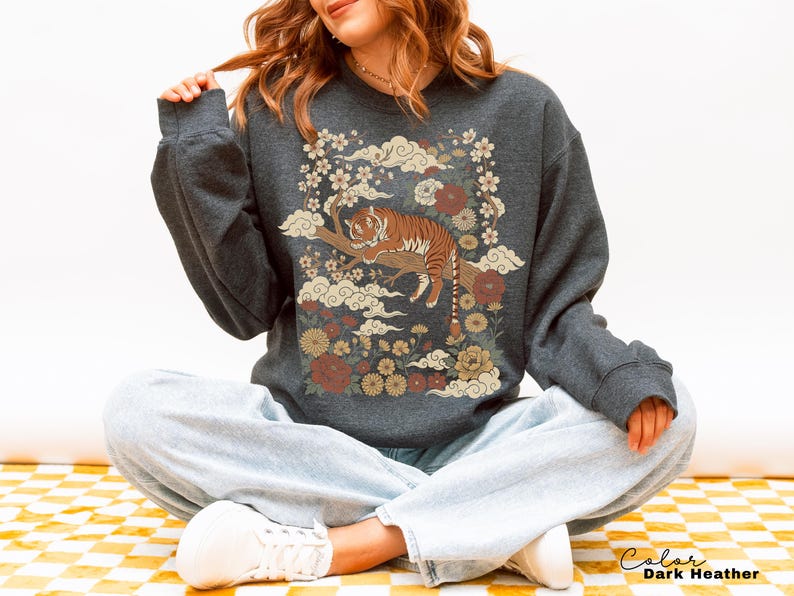 May include: Dark heather gray sweatshirt featuring a tiger resting on a branch, surrounded by flowers and clouds. The design includes shades of brown, cream, and red. The text "Color Dark Heather" is at the bottom right.