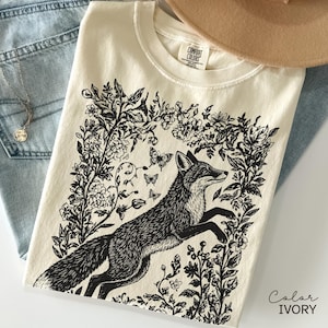 Pu&ograve; includere: T-shirt color avorio con un'illustrazione in bianco e nero di una volpe che salta attraverso fiori e farfalle. La maglietta &egrave; piegata su jeans blu. Un cappello beige &egrave; visibile sullo sfondo. Il testo "Color IVORY" &egrave; visibile.