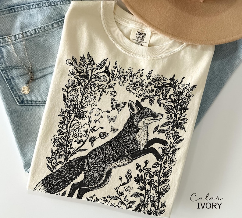 Pu&ograve; includere: T-shirt color avorio con un'illustrazione in bianco e nero di una volpe che salta attraverso fiori e farfalle. La maglietta &egrave; piegata su jeans blu. Un cappello beige &egrave; visibile sullo sfondo. Il testo "Color IVORY" &egrave; visibile.