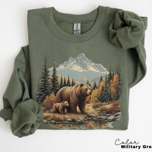 May include: Olive green sweatshirt featuring a mountain and forest scene with a mother bear and cub. The graphic includes a mountain range, trees, and a body of water. The text "Color Military Green" is at the bottom.