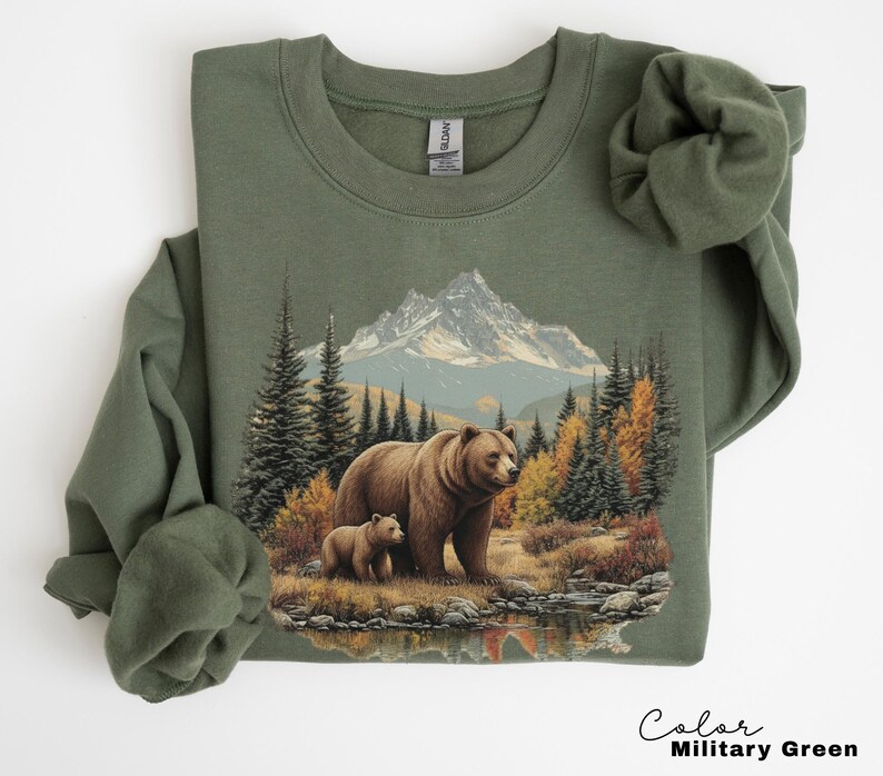 May include: Olive green sweatshirt featuring a mountain and forest scene with a mother bear and cub. The graphic includes a mountain range, trees, and a body of water. The text "Color Military Green" is at the bottom.
