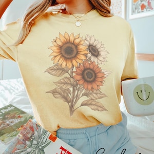 May include: A yellow t-shirt featuring a graphic of three sunflowers in shades of brown, orange, and white. The text "Color Banana" is written in cursive at the bottom of the image.