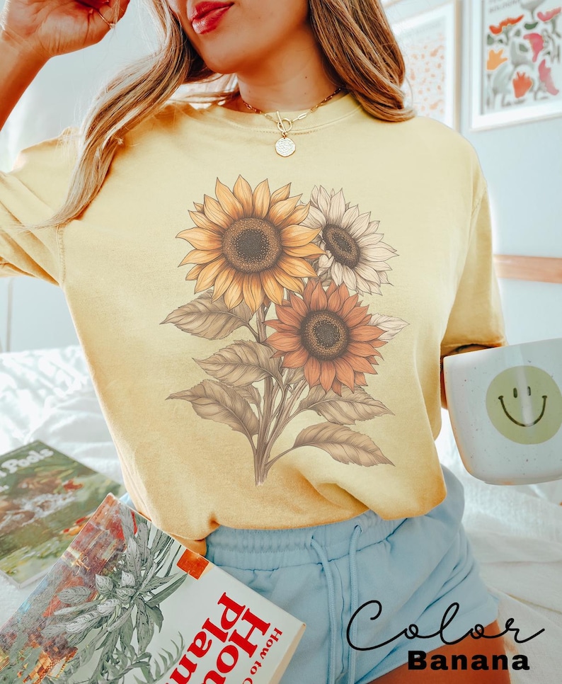 May include: A yellow t-shirt featuring a graphic of three sunflowers in shades of brown, orange, and white. The text "Color Banana" is written in cursive at the bottom of the image.