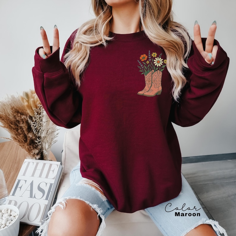 Puede incluir: Sudadera granate con un gr&aacute;fico de una bota de vaquero llena de flores coloridas. El texto "Color Maroon" est&aacute; escrito en cursiva. La sudadera tiene mangas largas y cuello redondo.