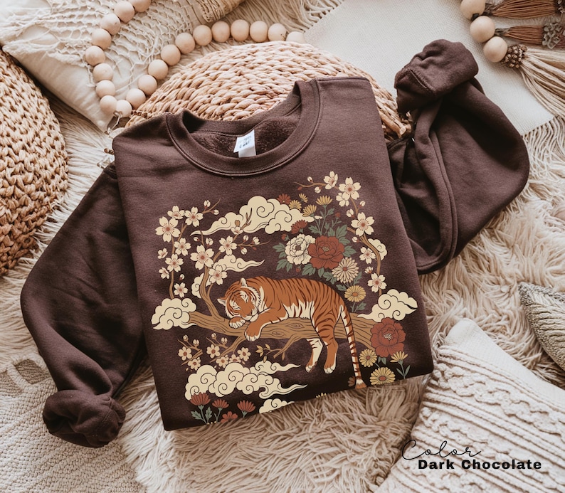 May include: Dark chocolate-colored sweatshirt featuring a tiger resting on a tree branch, surrounded by flowers and clouds. The design includes cream, brown, and red tones. The text "Color Dark Chocolate" is visible.