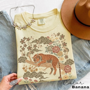 May include: A light yellow t-shirt with a graphic of a tiger resting on a tree branch, surrounded by flowers and clouds. The shirt is labeled "Color Banana". A necklace and a brown hat are also visible.