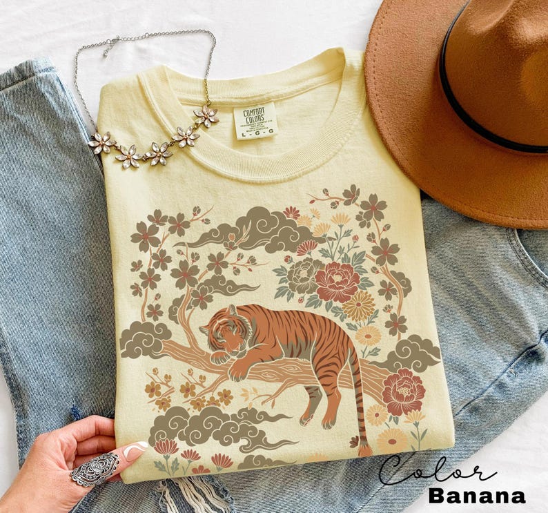 May include: A light yellow t-shirt with a graphic of a tiger resting on a tree branch, surrounded by flowers and clouds. The shirt is labeled "Color Banana". A necklace and a brown hat are also visible.