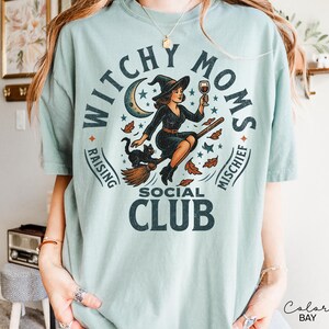 Puede incluir: Camiseta azul claro con un dise&ntilde;o circular de una bruja en una escoba, sosteniendo una copa de vino, con un gato negro. El texto dice "Witchy Moms Social Club, Raising Mischief."