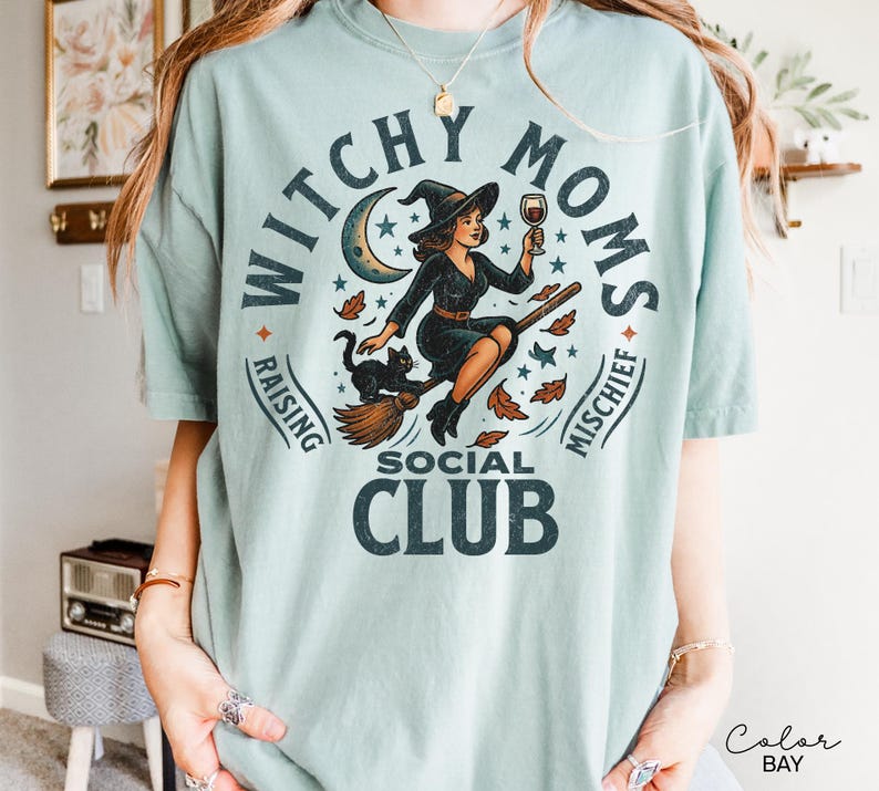 Puede incluir: Camiseta azul claro con un dise&ntilde;o circular de una bruja en una escoba, sosteniendo una copa de vino, con un gato negro. El texto dice "Witchy Moms Social Club, Raising Mischief."