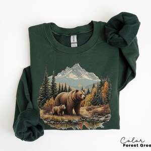 May include: Forest green sweatshirt featuring a detailed graphic of a mother bear and cub in a forest setting with a mountain backdrop. The design includes trees, water, and autumn foliage. The sweatshirt is a crew neck style.
