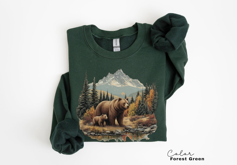 May include: Forest green sweatshirt featuring a detailed graphic of a mother bear and cub in a forest setting with a mountain backdrop. The design includes trees, water, and autumn foliage. The sweatshirt is a crew neck style.