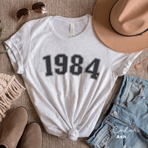 May include: A heather grey t-shirt with the year 1984 printed in black on the front. The shirt is styled with a brown hat, denim shorts, and brown boots.