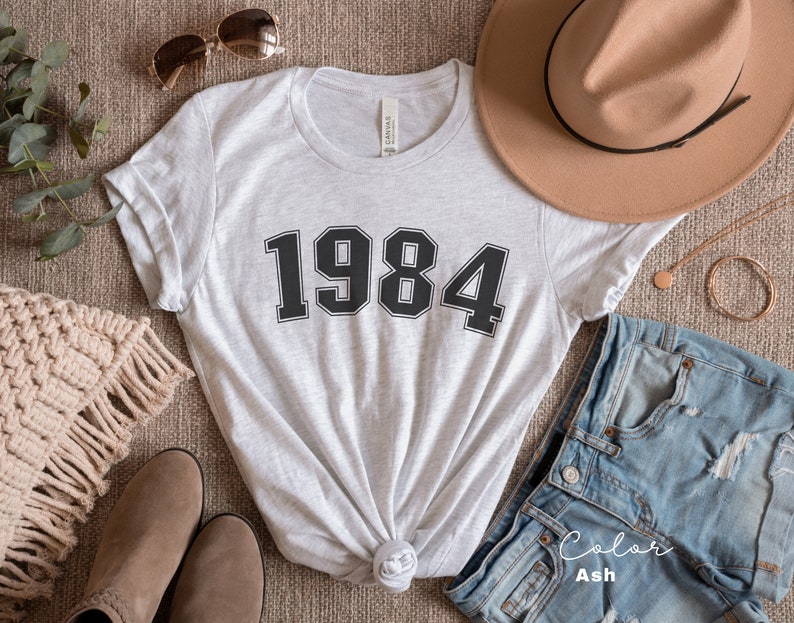 May include: A heather grey t-shirt with the year 1984 printed in black on the front. The shirt is styled with a brown hat, denim shorts, and brown boots.