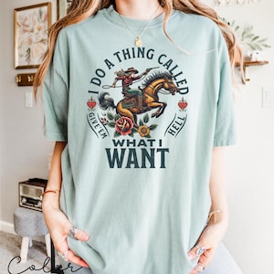 May include: A light blue t-shirt featuring a graphic of a woman riding a horse. The graphic is in a vintage style and includes the text "I do a thing called give 'em hell what I want".