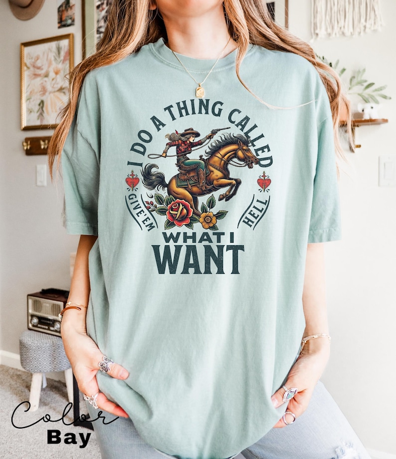 May include: A light blue t-shirt featuring a graphic of a woman riding a horse. The graphic is in a vintage style and includes the text "I do a thing called give 'em hell what I want".