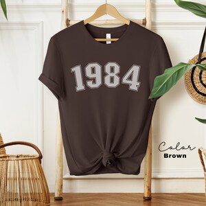 May include: A brown t-shirt with the year 1984 printed in white on the front. The shirt is knotted at the bottom and is hanging on a wooden ladder.