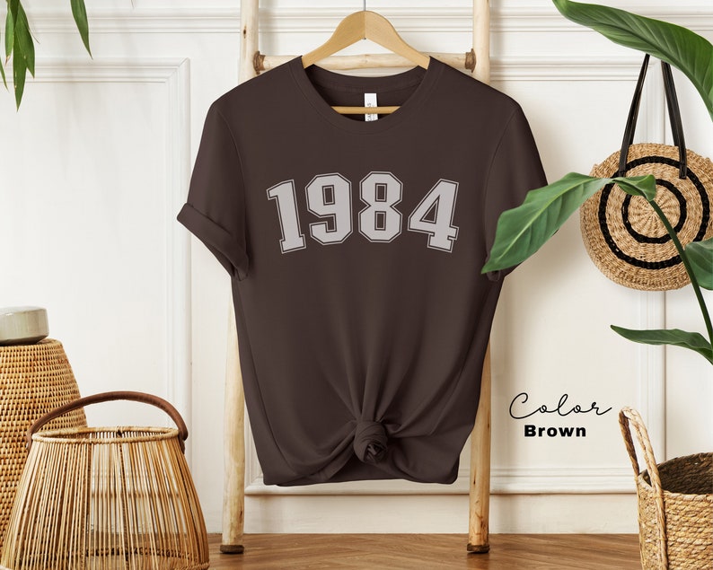 May include: A brown t-shirt with the year 1984 printed in white on the front. The shirt is knotted at the bottom and is hanging on a wooden ladder.