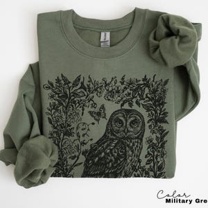 May include: Military green sweatshirt featuring a black illustration of an owl surrounded by floral elements and a butterfly. The sweatshirt has a crew neck and long sleeves. The text "Color Military Green" is at the bottom right.