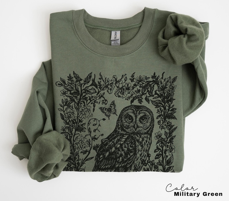 May include: Military green sweatshirt featuring a black illustration of an owl surrounded by floral elements and a butterfly. The sweatshirt has a crew neck and long sleeves. The text "Color Military Green" is at the bottom right.