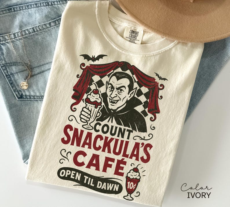 May include: Ivory-colored t-shirt featuring a graphic of Count Dracula holding a milkshake. The design includes the text "Count Snackula's Cafe" and "Open Til Dawn". The shirt is paired with blue jeans and a necklace.